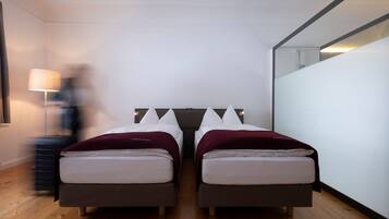 Een moderne hotelkamer met twee bedden, een houten vloer, een grote spiegel en een nachtkastje met een lamp.