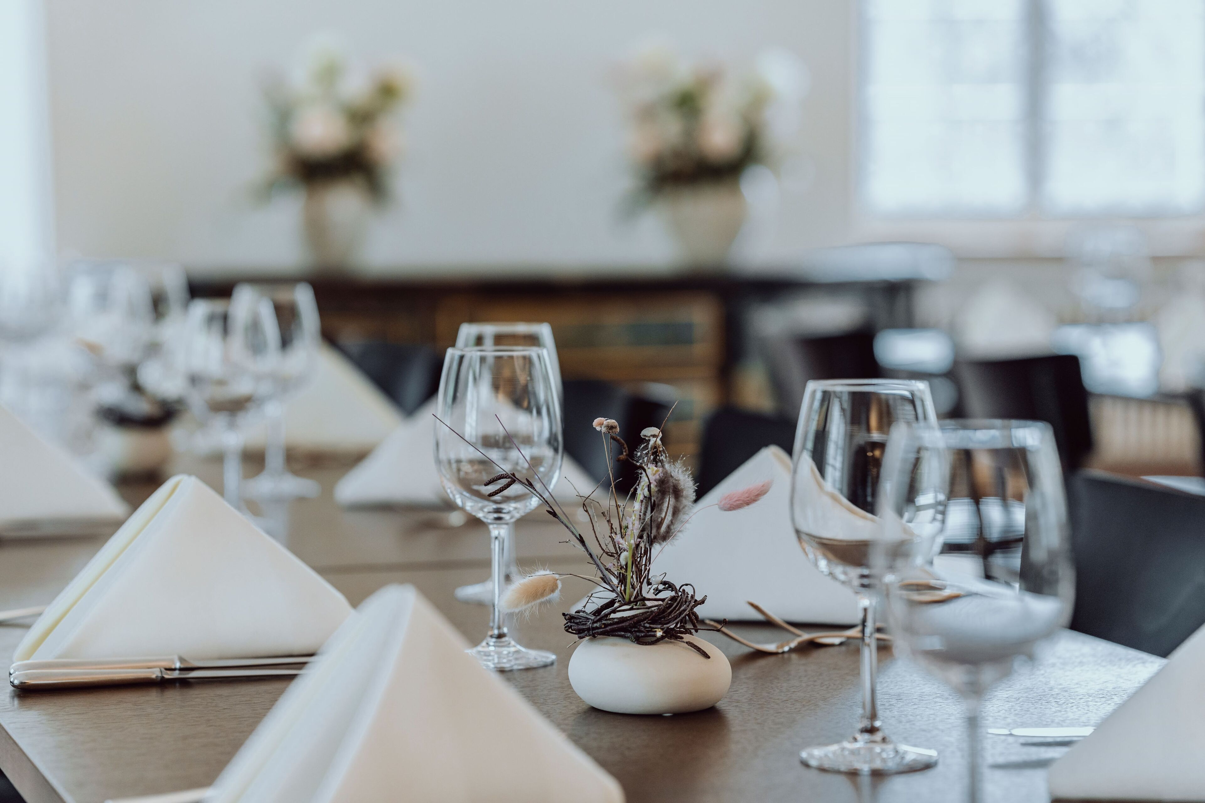 A dining table set with white napkins, wine glasses, and a centerpiece of dried flowers.