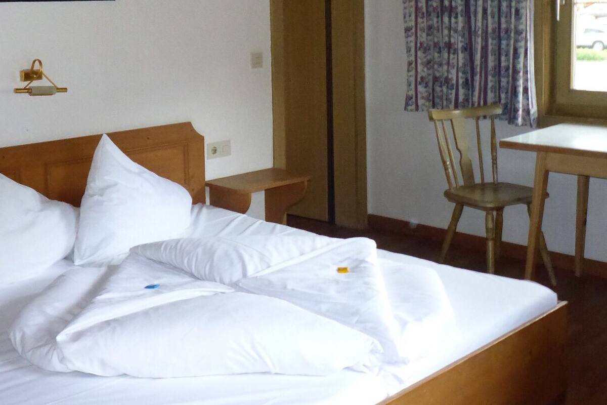 Un lit bien fait, avec du linge de lit blanc et des oreillers, une tête de lit en bois, une petite table de chevet en bois, une chaise et une table recouverte d’un rideau à motifs floraux.