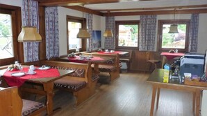 A dining area with wooden tables and benches, large windows with curtains, and hanging lamps. - Sportpension Penhab (Saalbach-Hinterglemm)