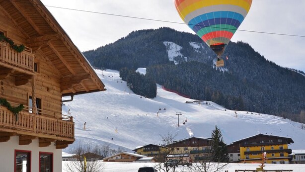 A wooden chalet with balconies, a ski slope in the background, and a hot air balloon. - Sportpension Penhab (Saalbach-Hinterglemm)