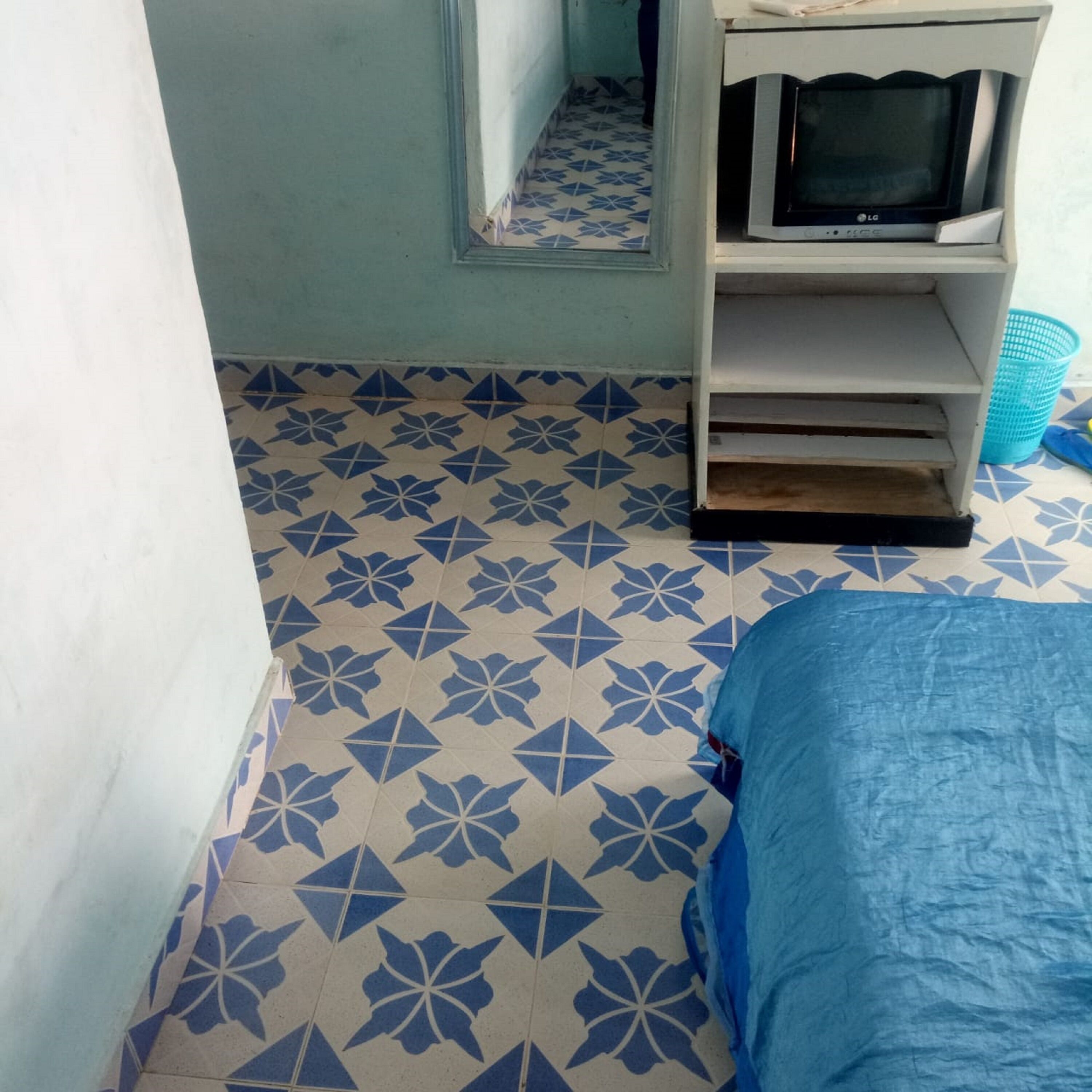 A room with a patterned floor, a bed with a blue cover, a shelf with a television, and a mirror on the wall.