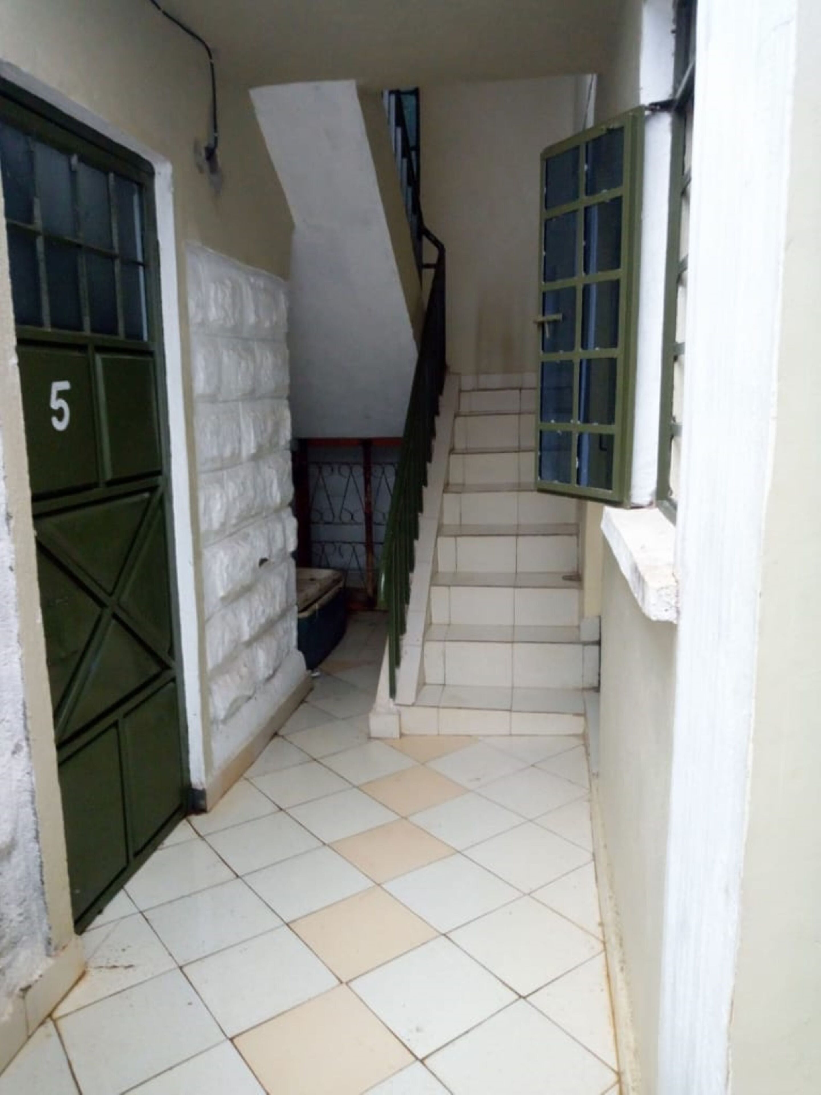 A narrow hallway with a tiled floor, a green door with a number, and a staircase with a green railing.
