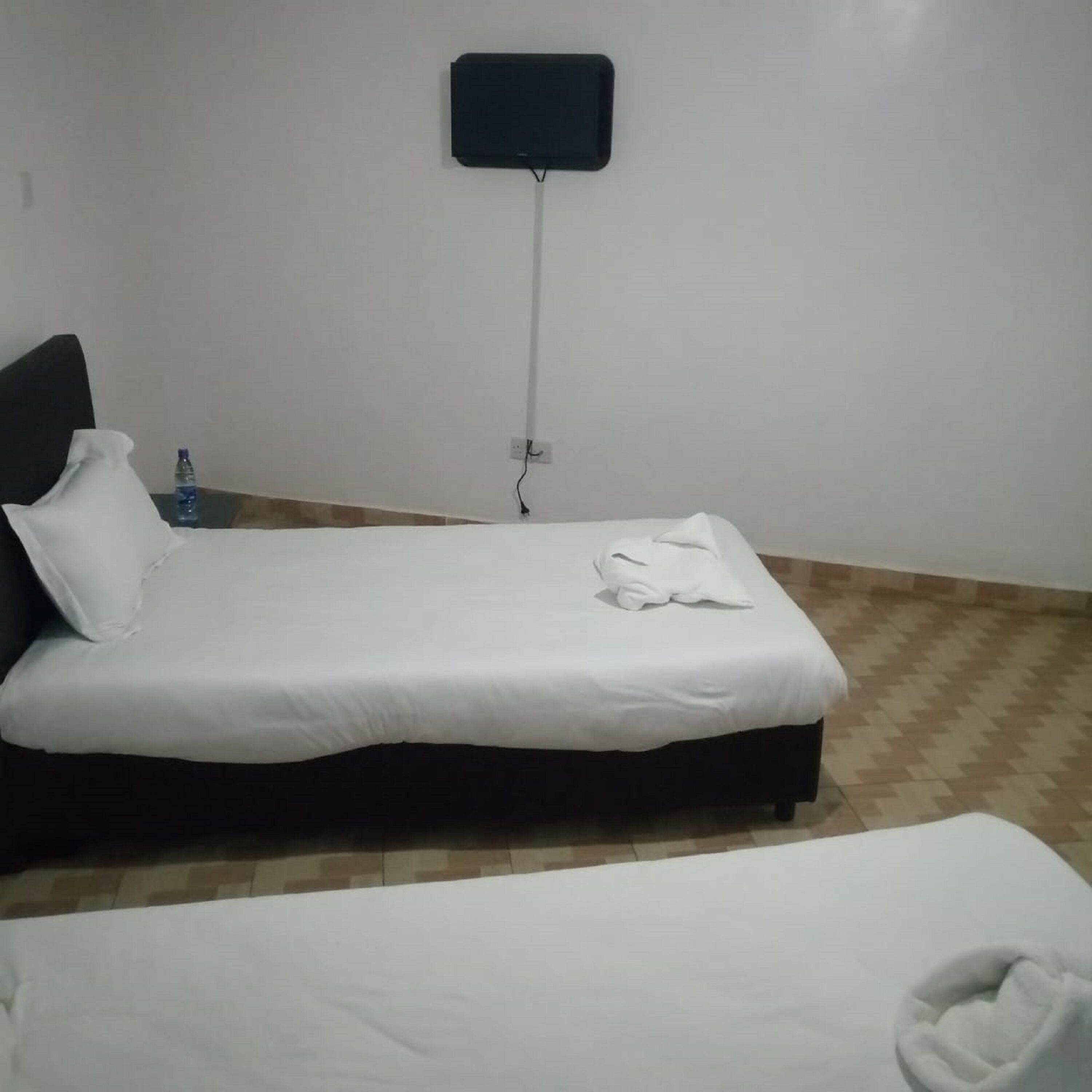 A single bed with a white mattress and pillow, a water bottle on the nightstand, and a power outlet on the wall.