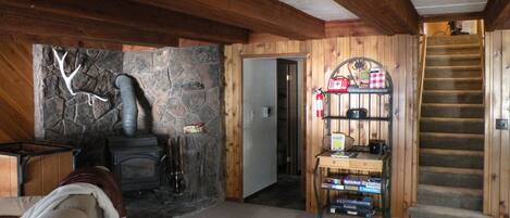 A cozy room with a stone wall, a fireplace, a wooden staircase, and a shelf with books and a fire extinguisher.