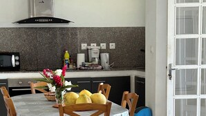 Une cuisine moderne dotée de plans de travail en granit, d’un four encastré et d’une machine à café. On y trouve un coin repas avec une table dressée pour quatre personnes, des chaises et un vase rempli de fleurs. Un canapé avec des coussins à rayures et un coussin décoratif est placé au premier plan.