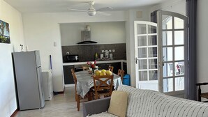 Un salon avec un canapé d’angle gris, une table à manger avec des chaises, un coin cuisine équipé d’un four à micro-ondes et d’une cuisinière, et une porte-fenêtre coulissante donnant sur un balcon.