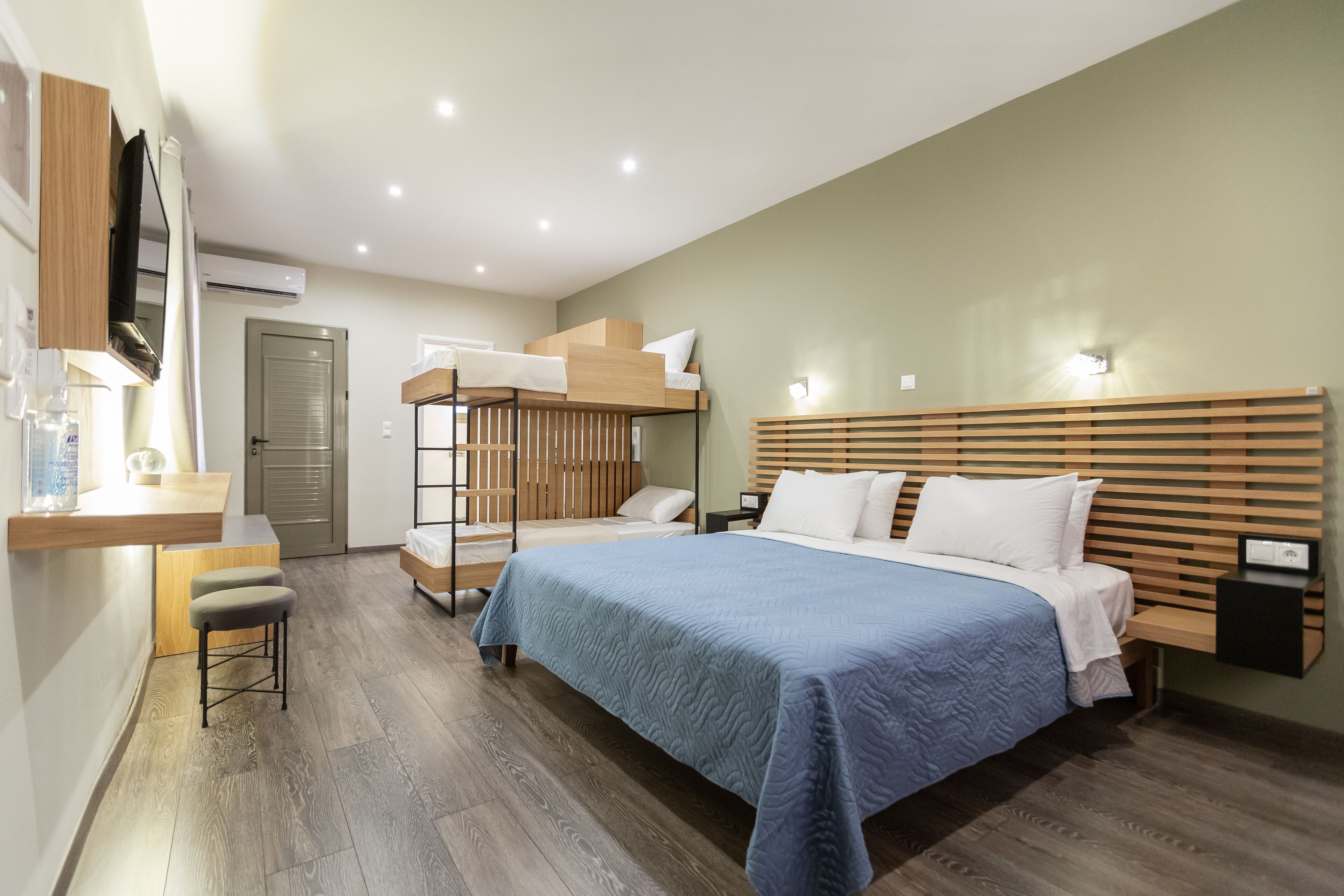 A modern hotel room with a bunk bed, a desk with a chair, a bedside table, and a flat-screen TV mounted on the wall.