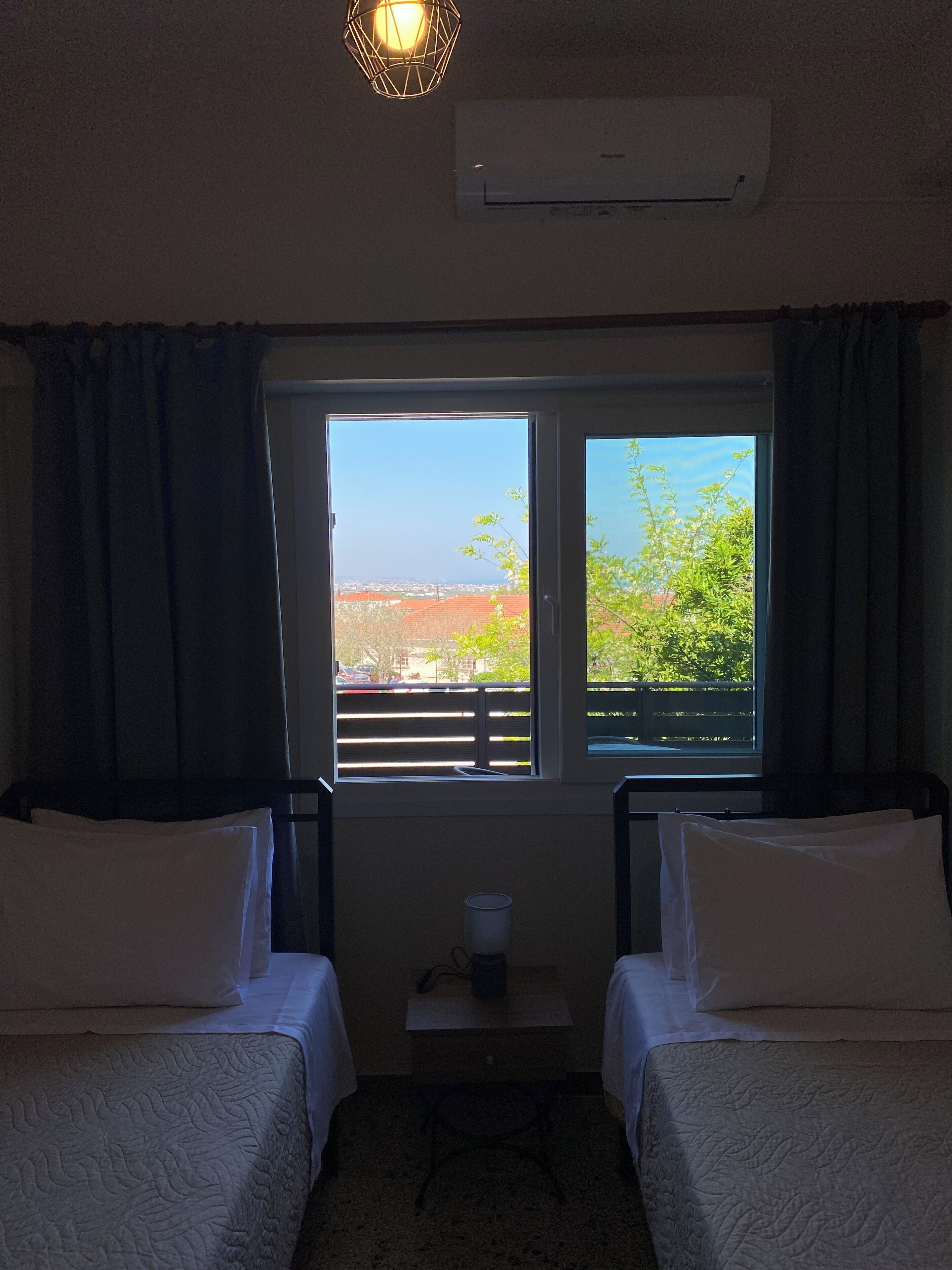 A hotel room with two beds, a window with a view, an air conditioning unit, and a small table with a cup.