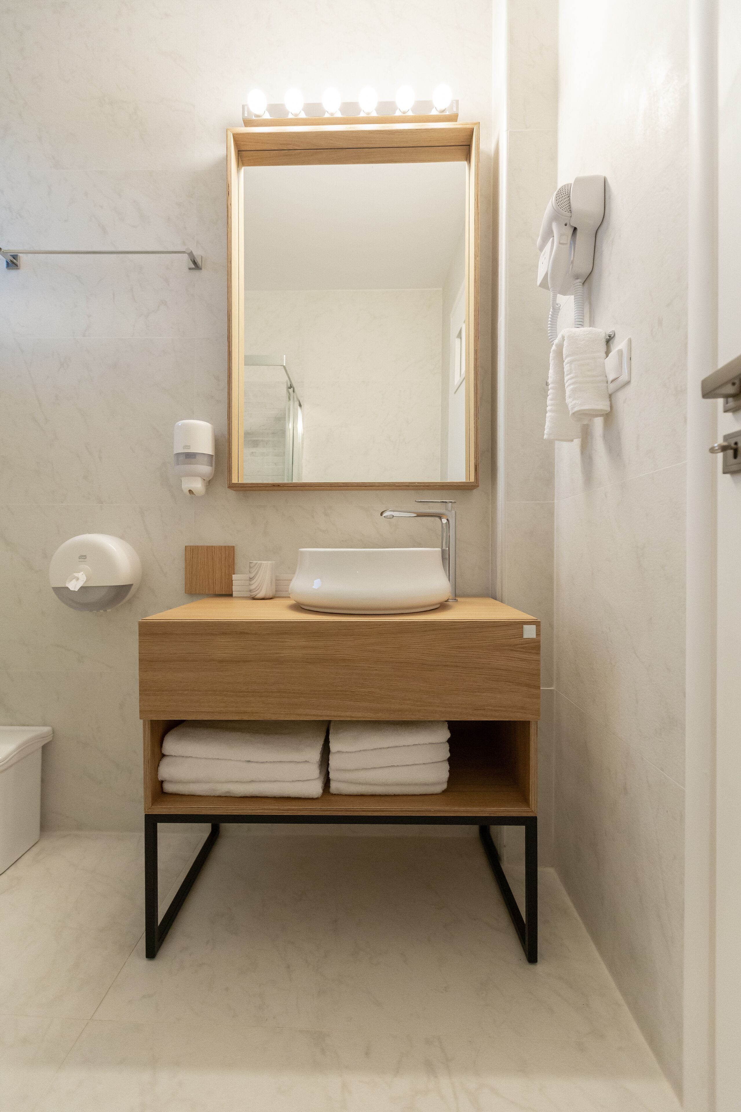 A modern bathroom with a wooden vanity, a large mirror with lights, a towel rack, and a hand dryer.