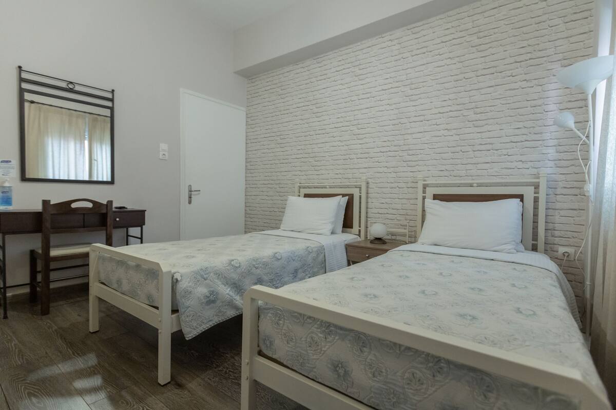 Ein Zimmer mit zwei Einzelbetten, einem Schreibtisch mit Stuhl, einem Spiegel und einer weißen Ziegelwand.