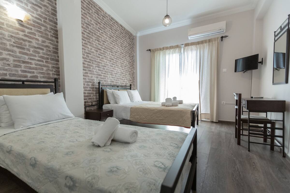 Ein Hotelzimmer mit Backsteinwand, einem Bett mit gemustertem Überwurf, einem weiteren Bett, einem Schreibtisch und einem an der Wand befestigten Fernseher.