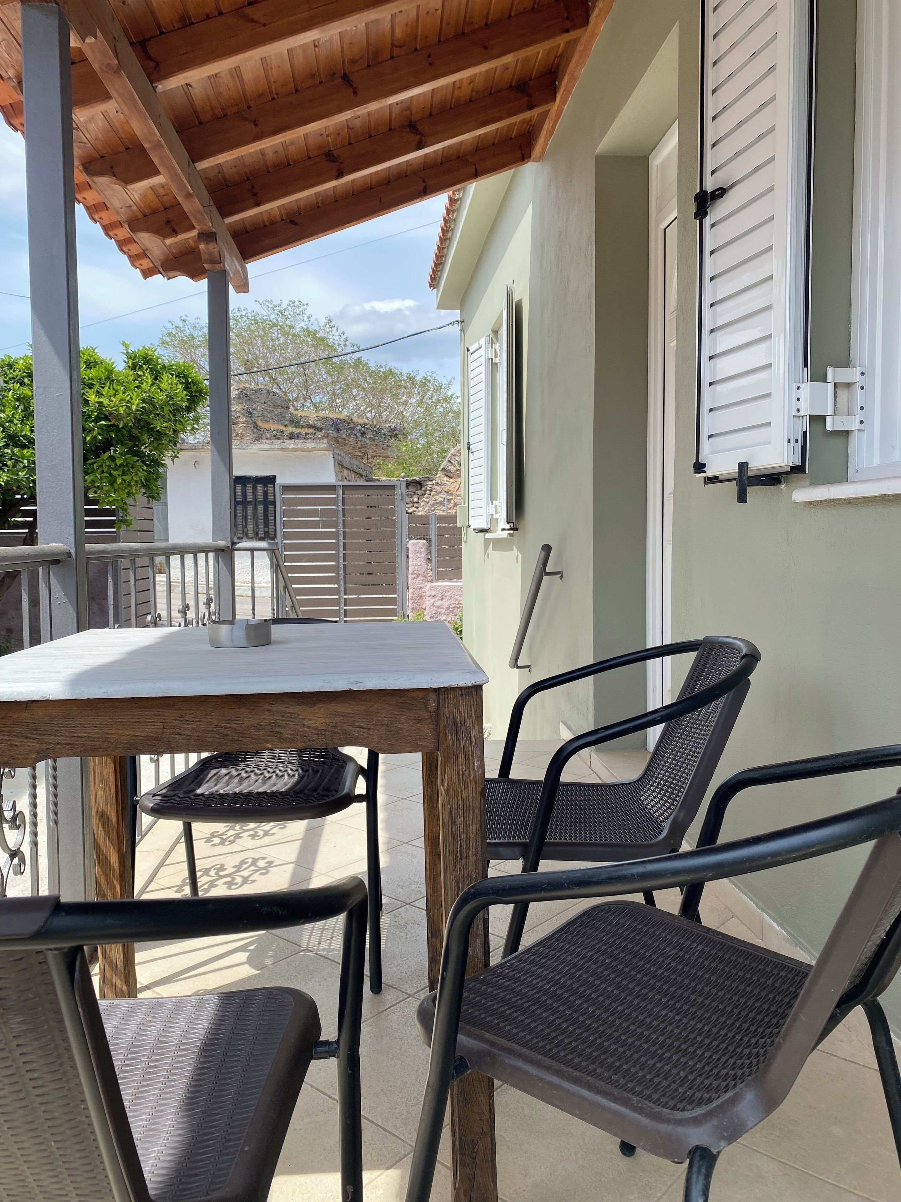 Une terrasse extérieure couverte, avec une table et des chaises, offrant une vue sur les environs, et un bâtiment doté de volets.