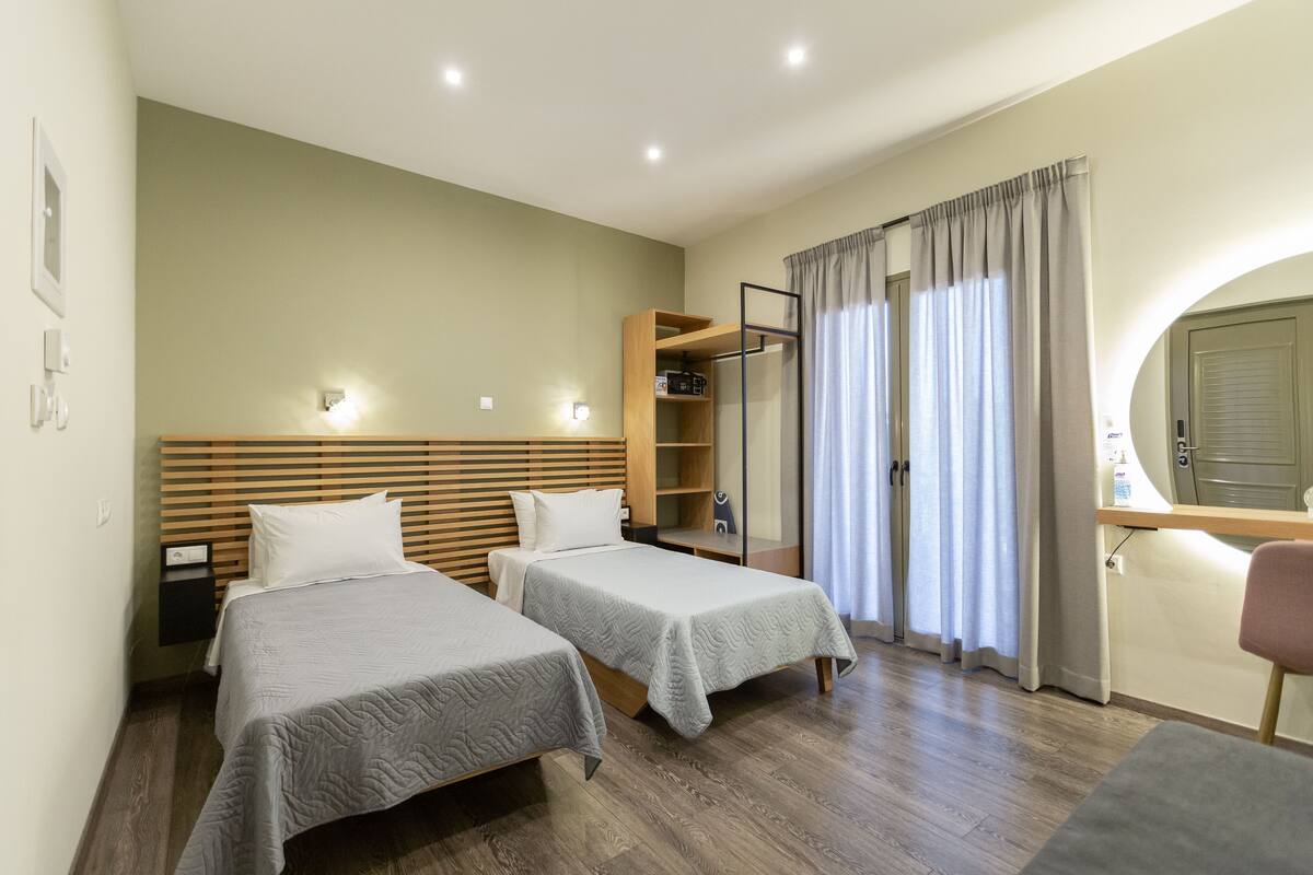 Ein Hotelzimmer mit zwei Betten, einem Holzkopfteil, Nachttischen, einem Kleiderschrank, einem Spiegel und einem Fenster mit Vorhängen.