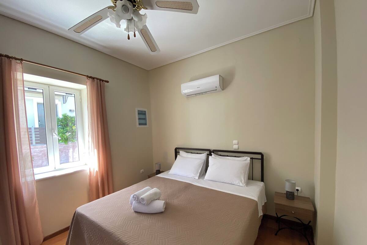 Ein Hotelzimmer mit Bett, Deckenventilator, Klimaanlage, einem Fenster mit Vorhängen und einem kleinen Nachttisch mit Lampe.