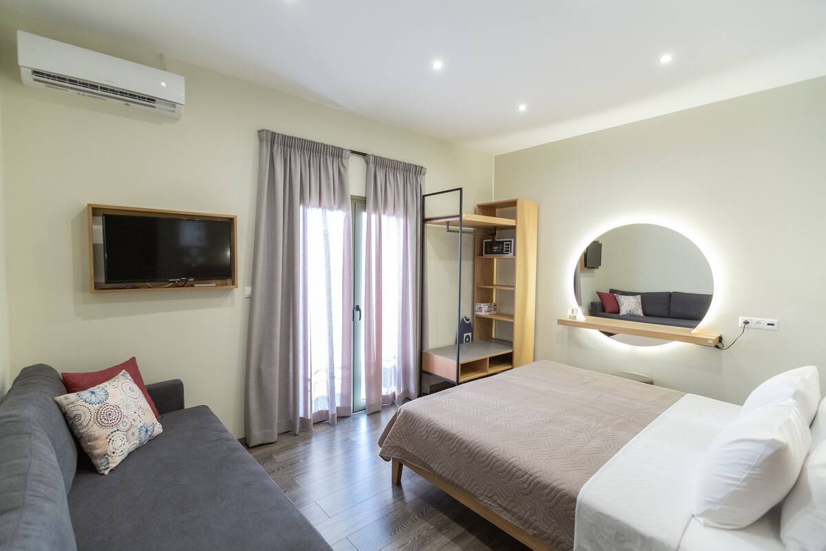Ein modernes Hotelzimmer mit Bett, Sofa, Fernseher, Kleiderschrank und einem runden Spiegel.