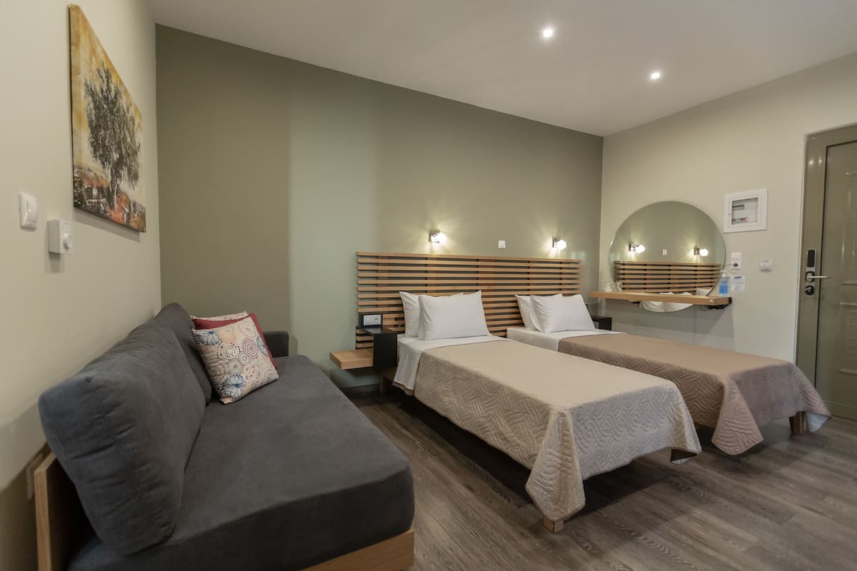 Ein Hotelzimmer mit einer Couch, zwei Einzelbetten, einem Holzkopfende, Wandleuchten, einem Spiegel und einer Tür.
