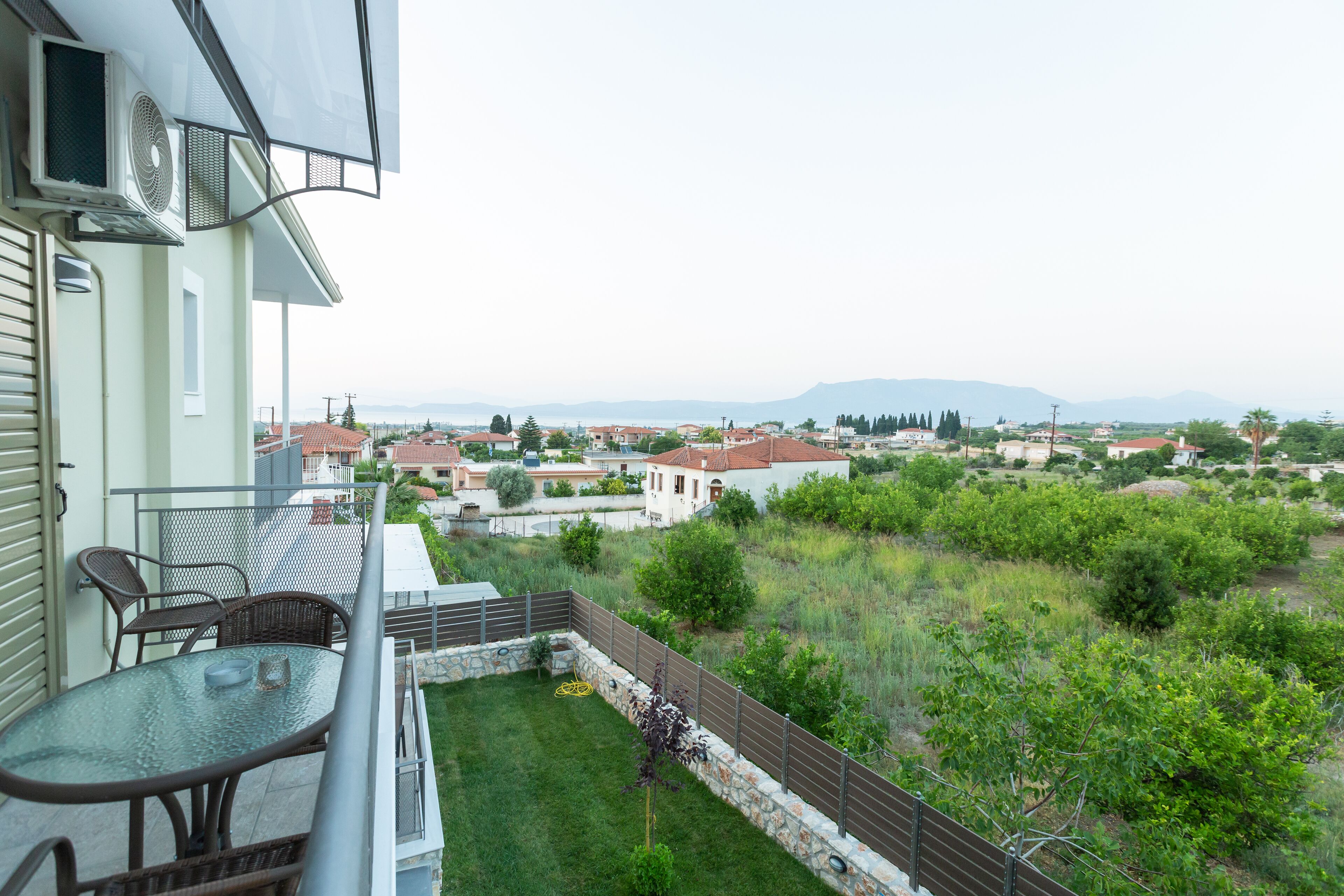 Un balcone con tavolo e sedie in vetro, affacciato su una zona residenziale con case e vegetazione.