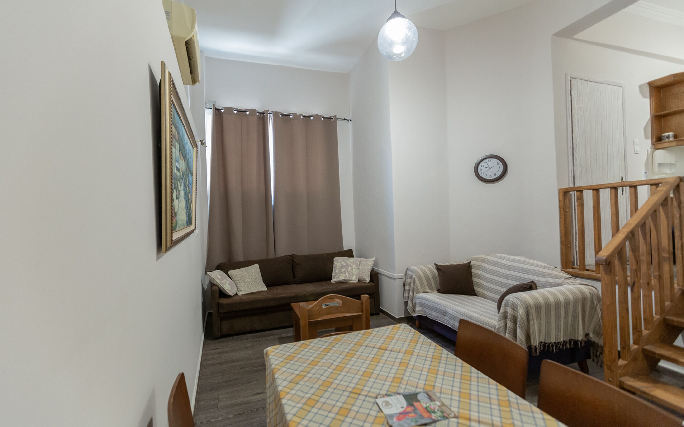 Una sala de estar pequeña y acogedora con un sofá, un sofá a rayas, una mesa de comedor con mantel a cuadros, una silla de madera y una escalera de madera, además de un reloj en la pared.