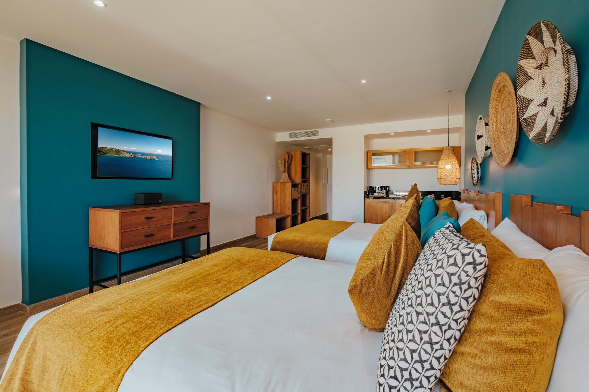 A hotel room with a large bed, a TV, a wooden dresser, and a kitchen area visible through an open door.