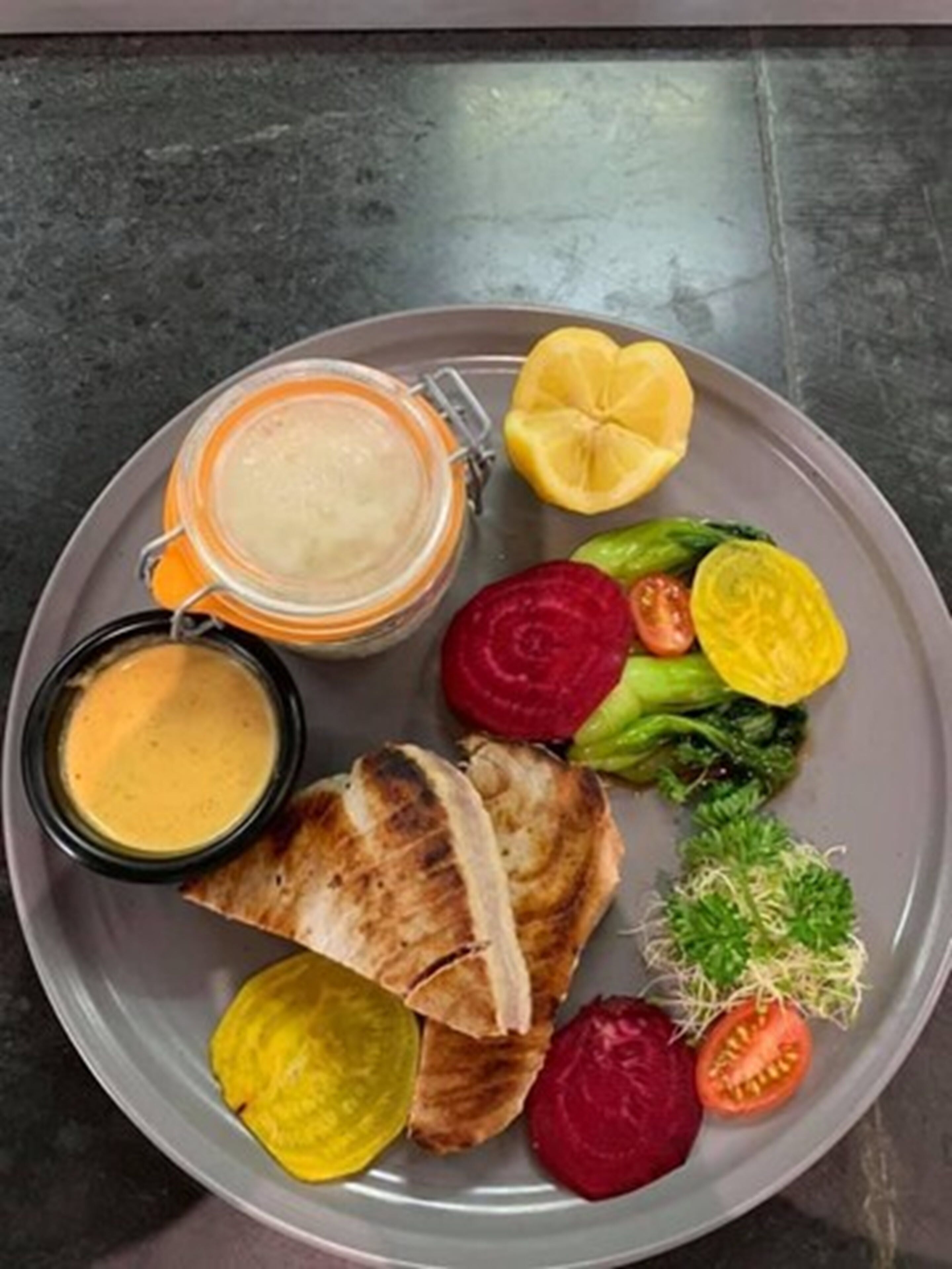 A plate with grilled meat, sliced vegetables, and two small containers of sauce.