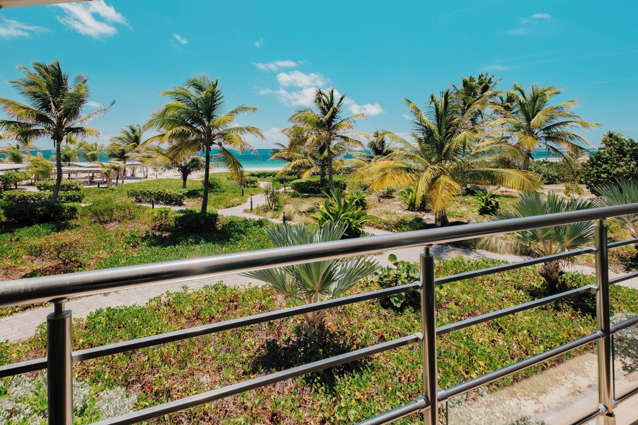 A balcony with a metal railing overlooking a landscaped area with palm trees and a beach in the background.