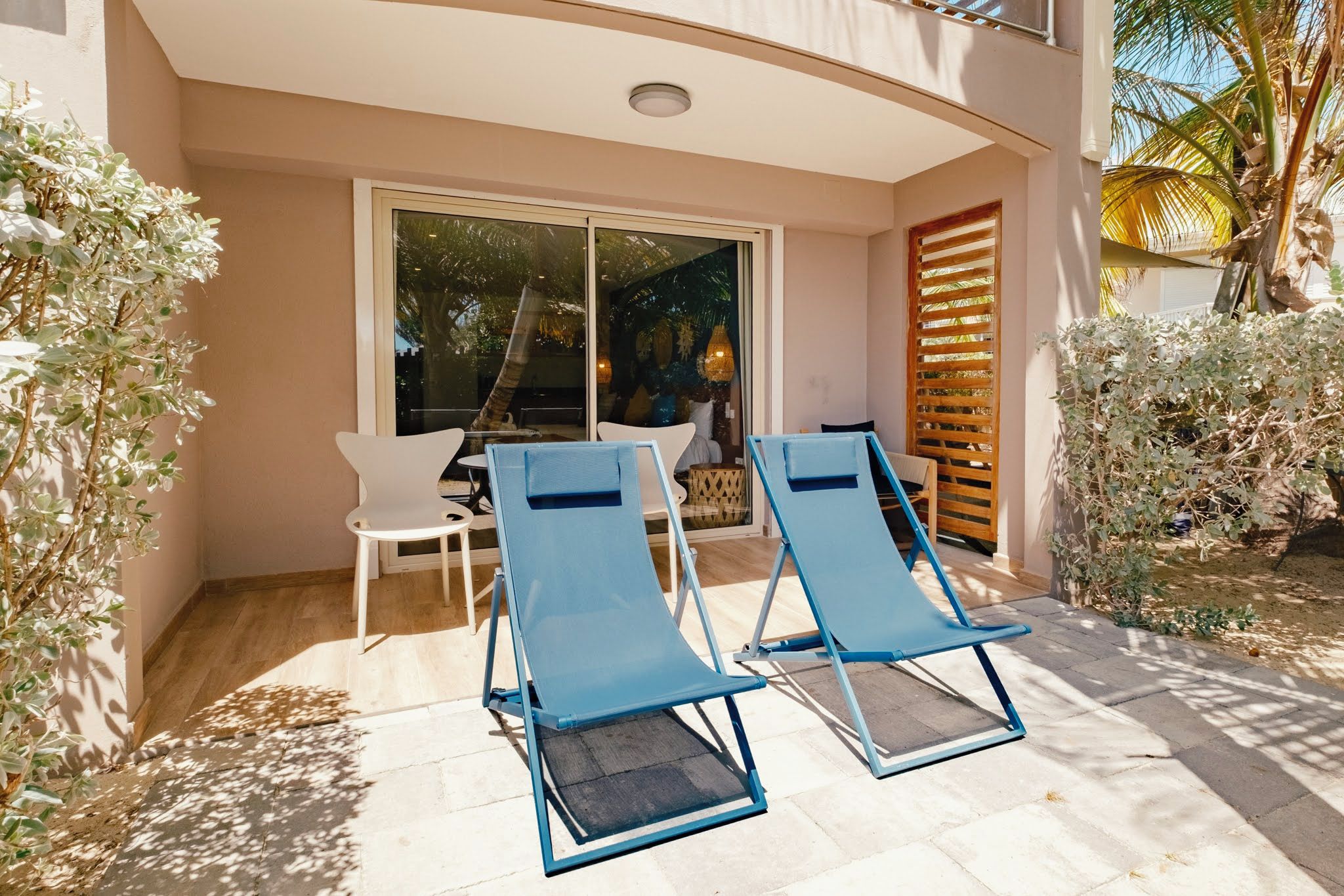 A patio with two blue lounge chairs and a white chair, a glass door leading to an indoor area, and a wooden fence.