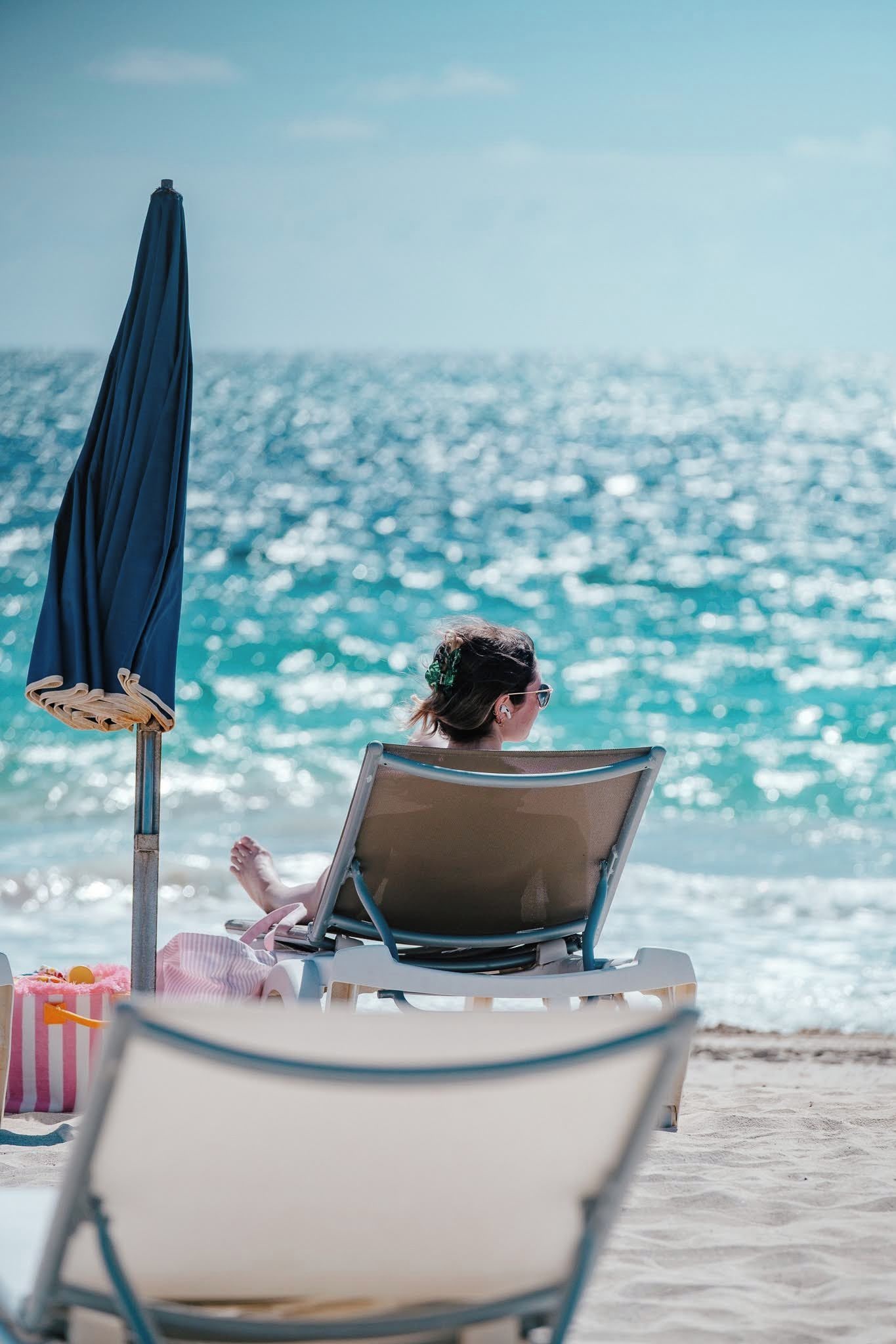 Eine Strandansicht mit einer Person, die auf einer Liege entspannt, einem blauen Sonnenschirm und einer gestreiften Strandtasche.