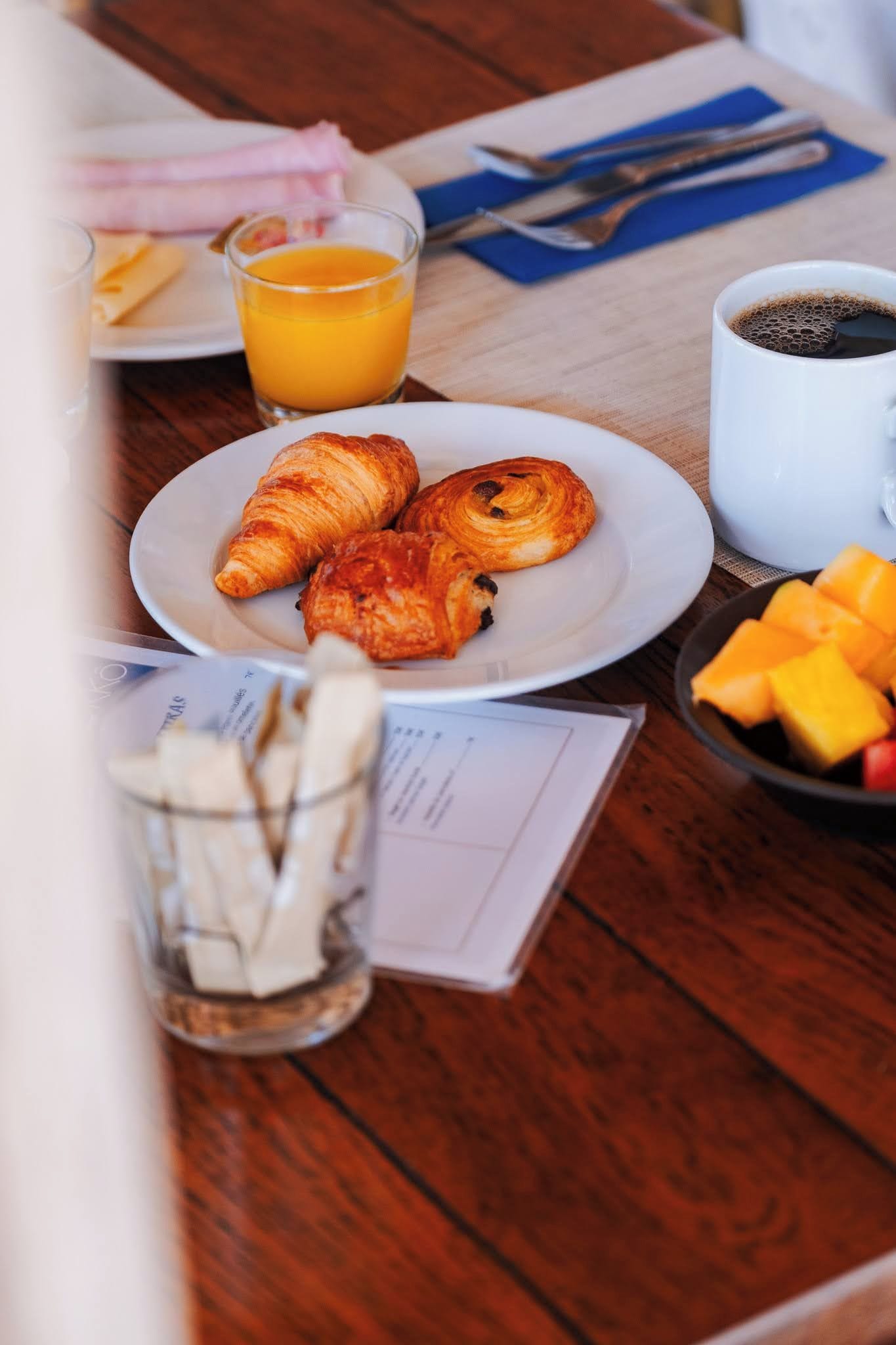 A breakfast setting with croissants, orange juice, coffee, and fruit on a wooden table.
