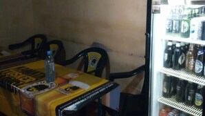 Interior de un restaurante con una mesa cubierta de material promocional, un refrigerador lleno de bebidas y sillas alrededor de la mesa.