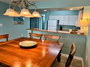 A dining area with a wooden table and chairs, a chandelier, and a kitchen with white cabinets and a refrigerator. - Blue Water Keyes-Oceanfront- Updated 2/2 -Multiple Pools-Lazy River-Family Fun (North Myrtle Beach)