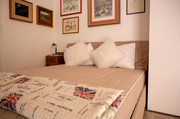 Un lit bien fait, recouvert d’une courtepointe, une table de chevet avec une lampe, et quatre tableaux encadrés accrochés au mur.