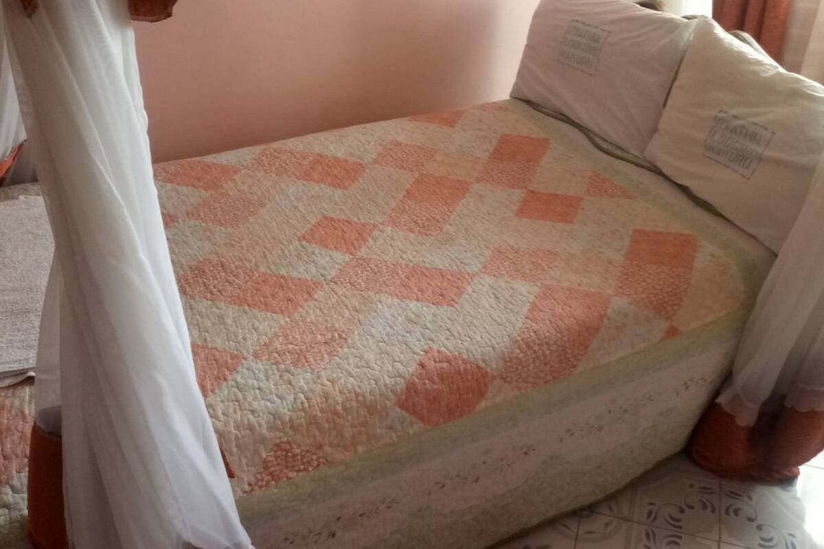 A bedroom with a checkered bedspread, pillows, a bed, a curtain, and a pair of slippers on the floor.