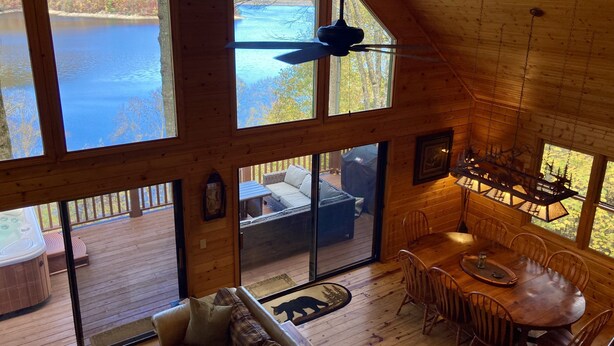 Una cabaña con amplias ventanas con vistas a un lago, un comedor con mesa y sillas de madera, y una zona de estar acogedora con sofá y jacuzzi.