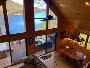 A cabin with large windows overlooking a lake, a dining area with a wooden table and chairs, and a cozy seating area with a sofa and a hot tub.