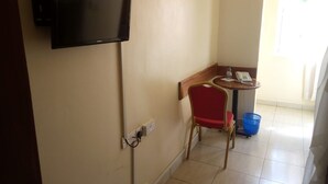 A hotel room with a flat-screen TV mounted on the wall, a small round table with a chair, a blue trash bin, and a telephone. - Bungoma Midtown Hotel (Bungoma)