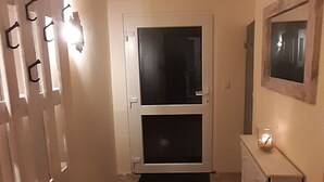 En smal gang med flisegulv, en dør med sort dørmåtte og et spejl på væggen.
