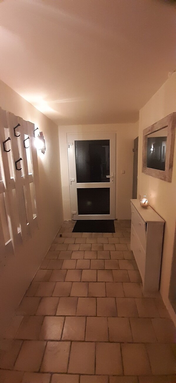A narrow hallway with a tiled floor, a door with a black mat, and a wall-mounted mirror.