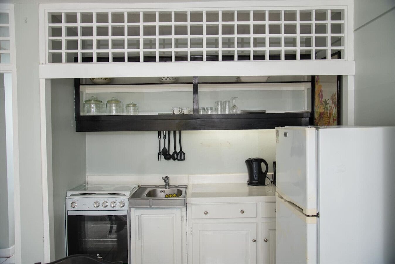 A compact kitchen with a white stove, sink, and refrigerator. Above the sink, there is a shelf with glass jars and a kettle.