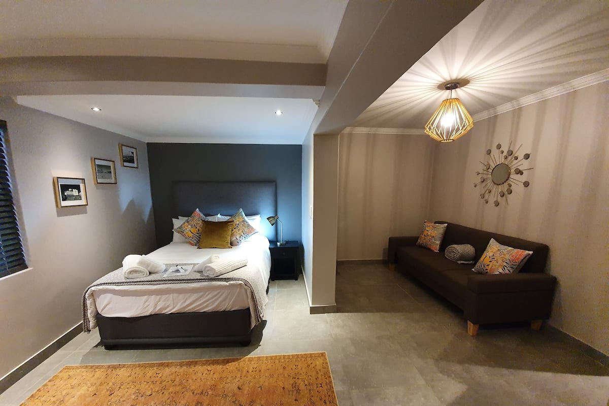 Un dormitorio moderno con una cama grande, un cabecero oscuro, una mesita de noche, un sofá y un elemento decorativo en la pared.