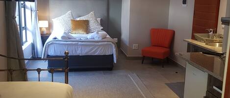 A bedroom with a bed, a red chair, a small kitchenette, and a ceiling light.