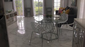Una habitación amplia con suelo de mármol, una mesa de vidrio con sillas, un sofá con cojines y ventanas grandes con cortinas.