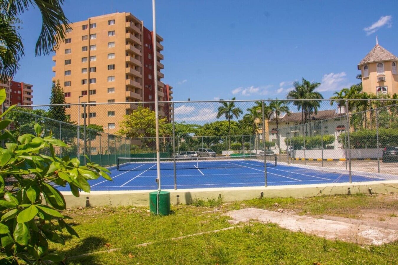 A tennis court surrounded by a chain-link fence, with buildings in the background.