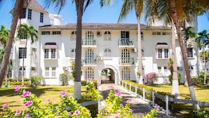 A white building with multiple windows and balconies, surrounded by palm trees and a garden with pink flowers. - Sunflower Serenade (Ocho Rios)