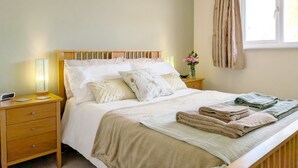 Una cama bien hecha con cabecera de madera, una mesita de noche con lámpara, y una ventana con cortinas.