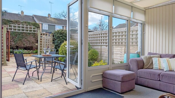 Un invernadero con puerta de cristal, una zona de estar con sofá y reposapiés, y un patio con mesa y sillas.