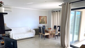 Un saló modern amb un sofà blanc, una taula de dinar de vidre amb cadirees, un televisió de pantalla plana i una finestra gran amb cortines.