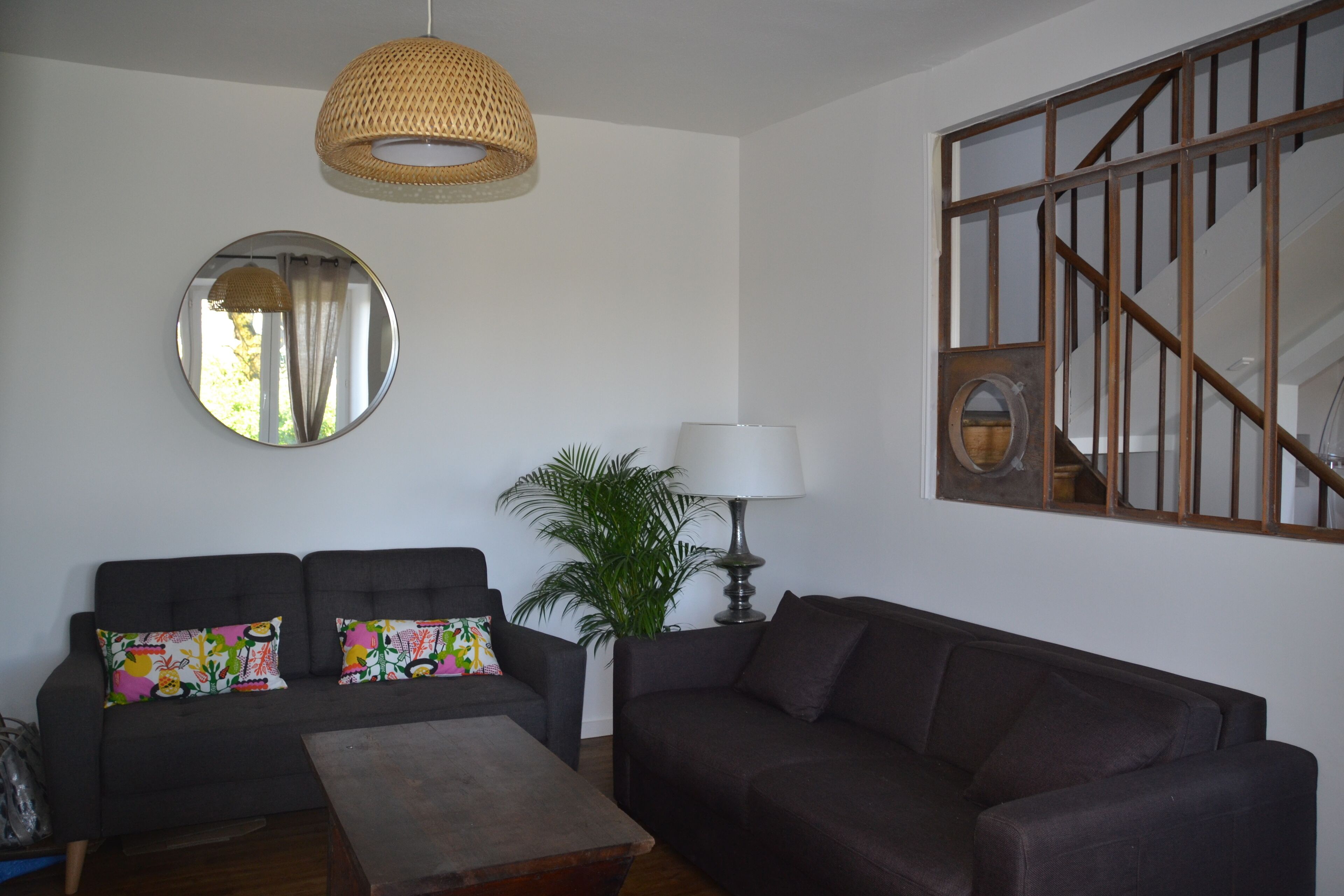 Un salon comprenant un canapé sombre, une table basse, un miroir rond, une lampe et une grande fenêtre donnant sur un escalier.