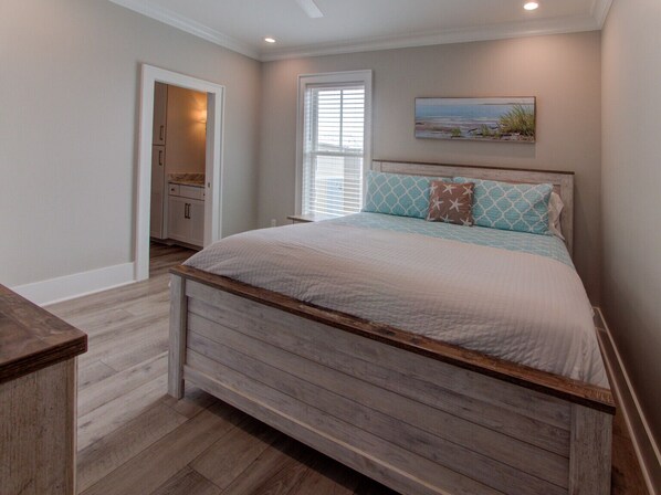 A bedroom with a wooden bed frame, a window with blinds, a painting on the wall, and a bathroom visible through an open door.