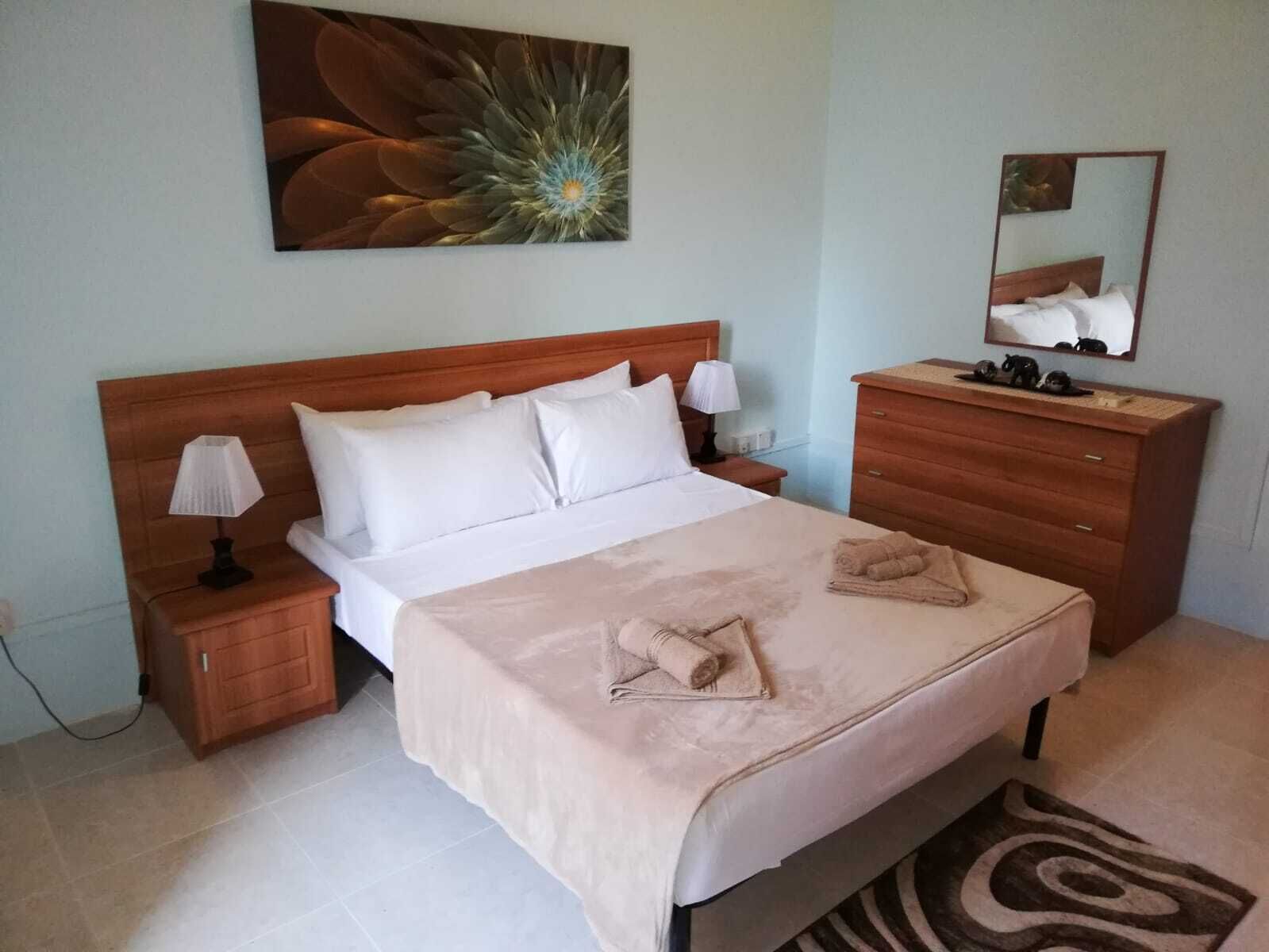 A neatly made bed with white linens, a wooden headboard, a bedside lamp, a wooden dresser, a mirror, and a decorative wall piece.
